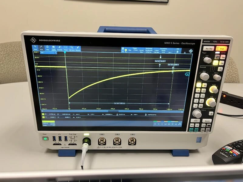 A Rohde and Schwarz MXO 5 Series oscilloscope on a table, showing a yellow trace that drops sharply and then curves back up toward its starting level.